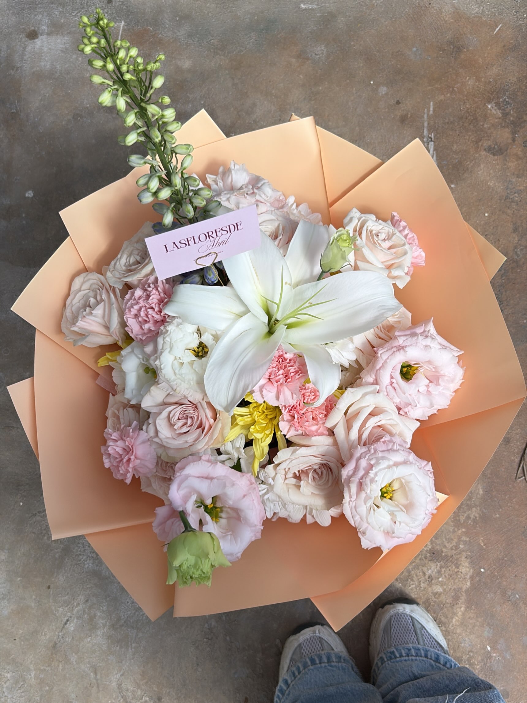 Ramo de Las Flores de Abril con lirio blanco, rosas y flores en tonos pastel, envuelto en papel durazno